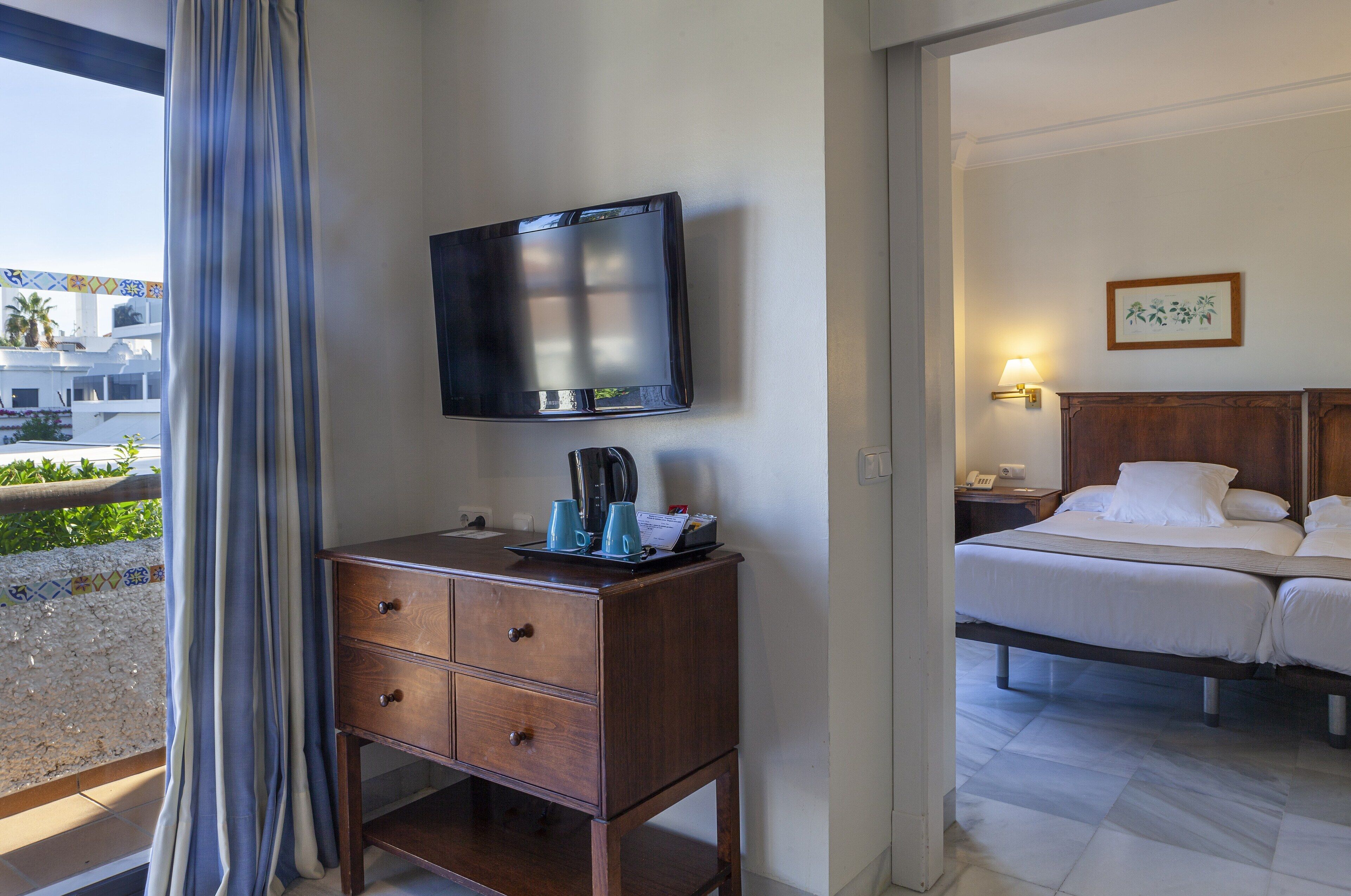 Habitación de hotel con cama, televisor montado en una cómoda de madera, mesita de noche con lámpara y vista al exterior.