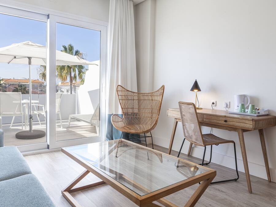 A modern room with a glass table, two wicker chairs, a wooden desk, and a view of the outdoor area with umbrellas and palm trees.