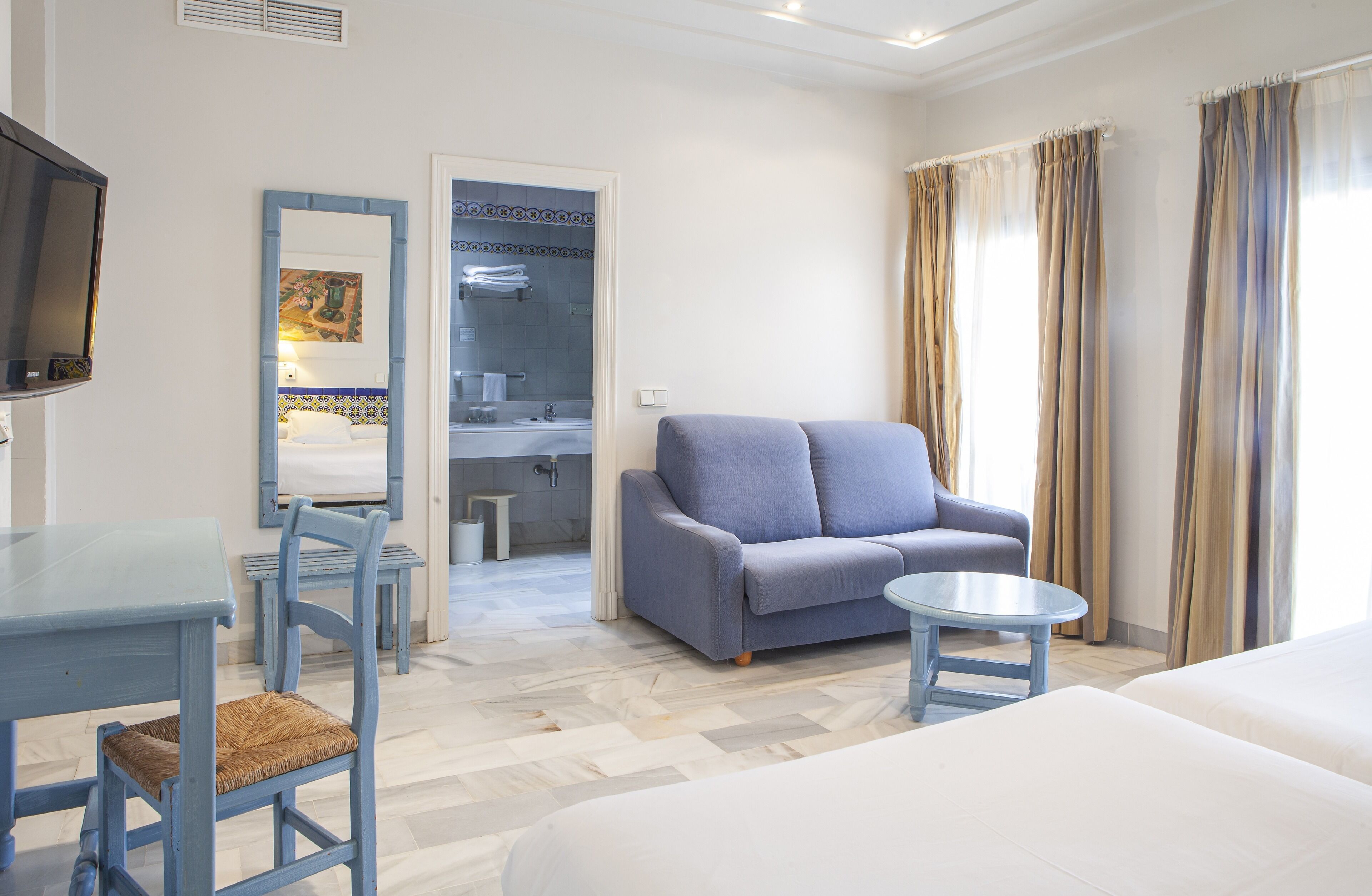 A hotel room with a bed, a sofa, a small table, a TV, a bathroom with a mirror, and a window with curtains.