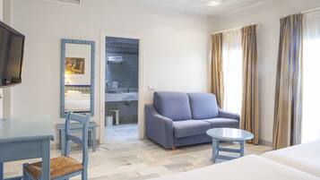A hotel room with a bed, a sofa, a small table, a TV, a bathroom with a mirror, and a window with curtains.