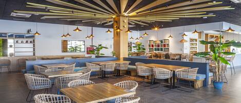 A modern restaurant interior with wooden tables and wicker chairs, a central wooden column, and pendant lighting.
