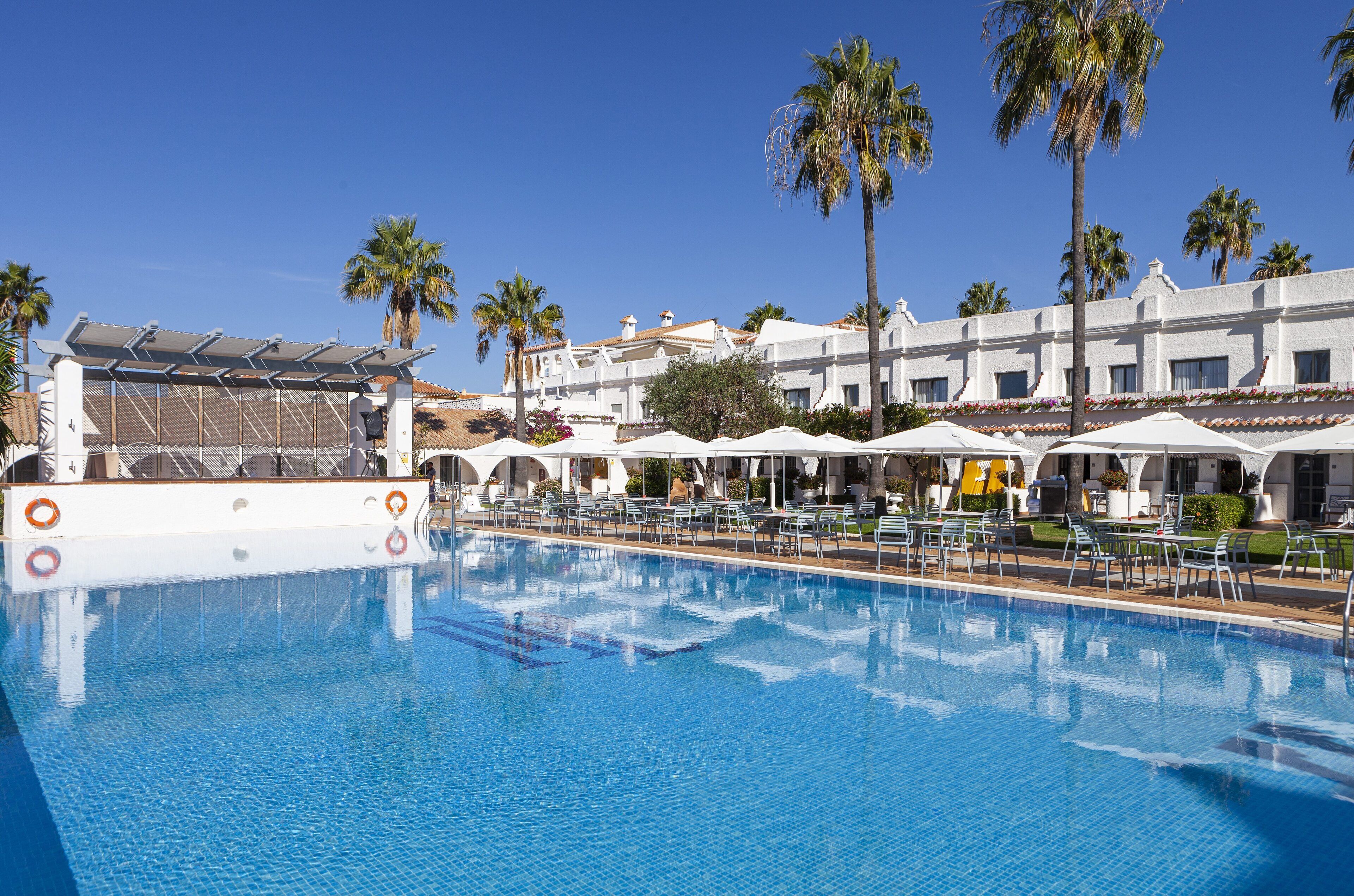 Una piscina con sillas de descanso y sombrillas, rodeada de palmeras y un edificio blanco con arcos.