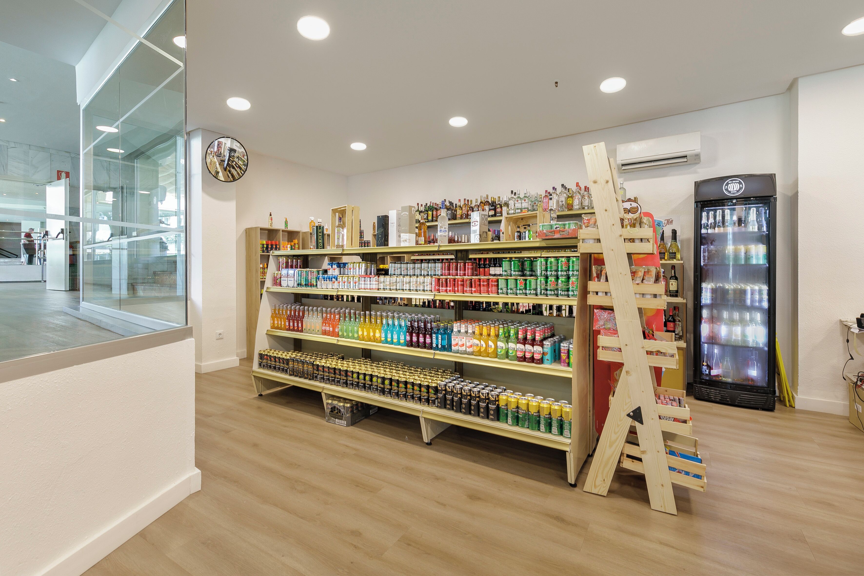 Interior de una tienda con estantes llenos de diversas bebidas, un refrigerador y una escalera de madera.