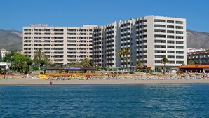 Un grand hôtel à plusieurs étages, avec une plage au premier plan et des montagnes en arrière-plan.