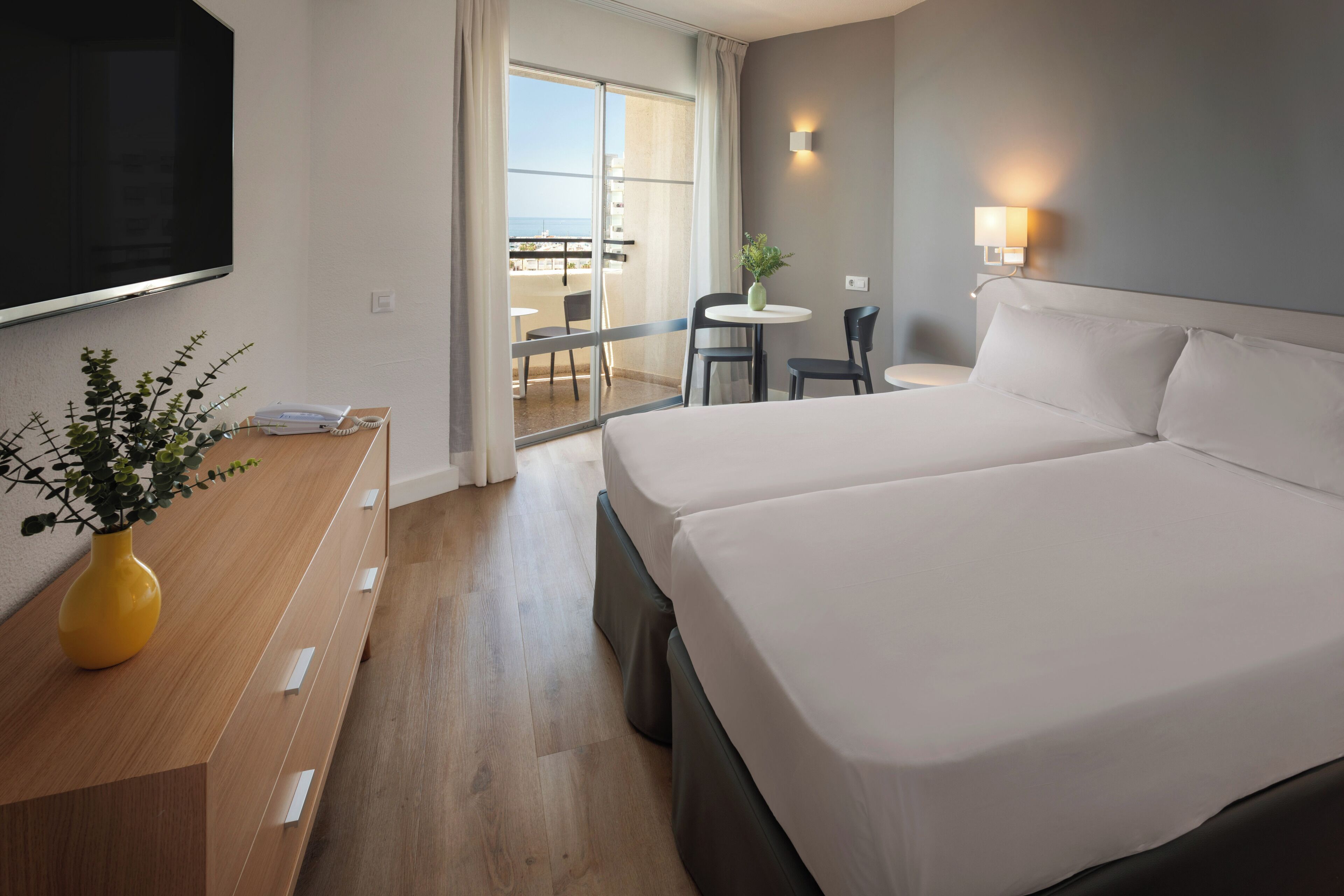 Habitación de hotel con una cama grande, televisor montado en la pared, escritorio con un jarrón de flores y balcón con mesa y sillas.