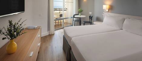 Habitación de hotel con una cama grande, televisor montado en la pared, escritorio con un jarrón de flores y balcón con mesa y sillas.