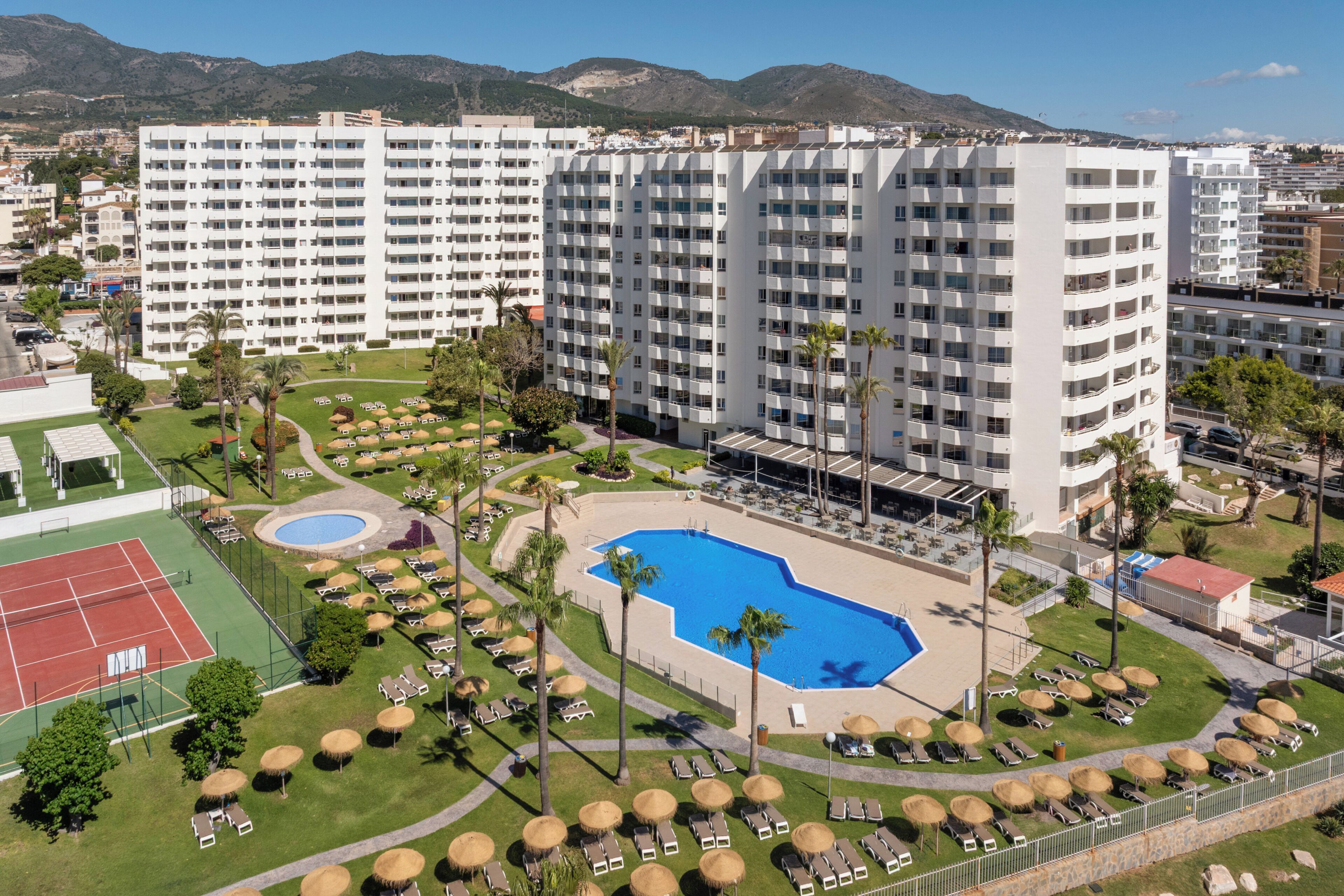 A hotel with multiple floors, a swimming pool, tennis courts, and sun loungers.