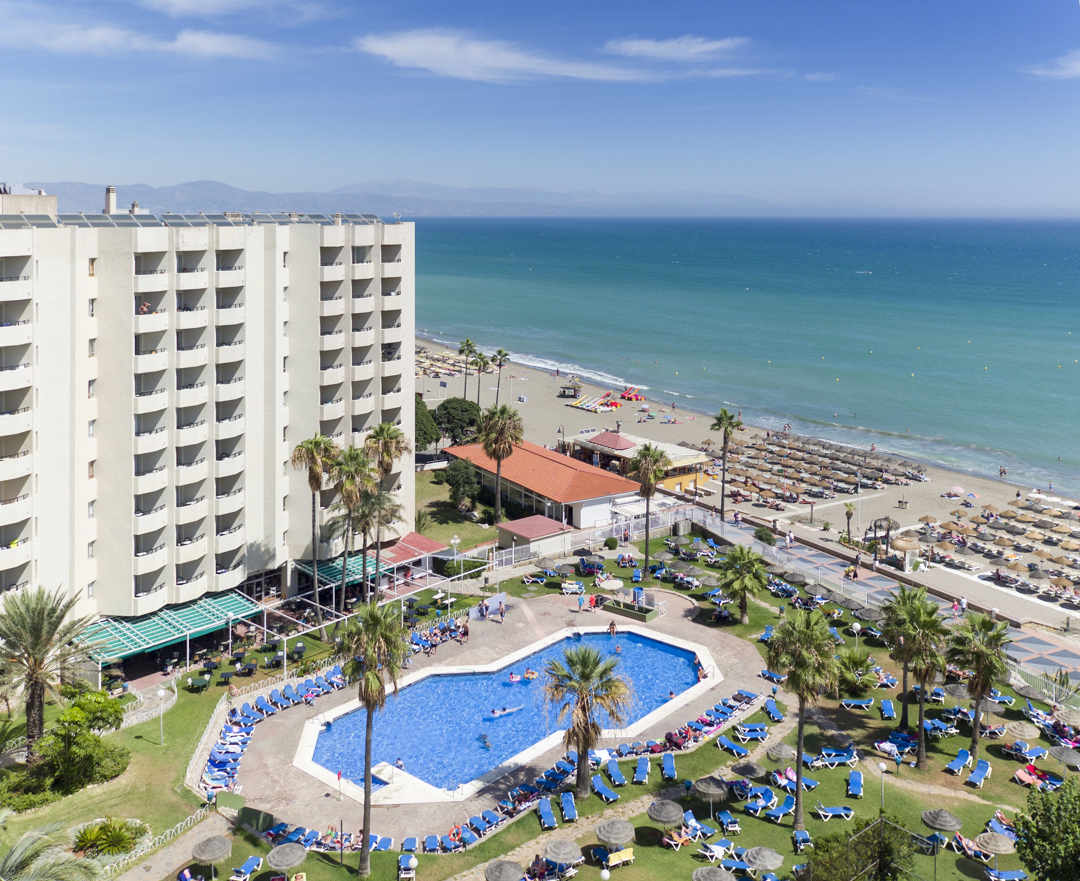 Ein Hotel mit Pool, umgeben von Liegestühlen und Palmen, mit Blick auf einen Strand und den Ozean.