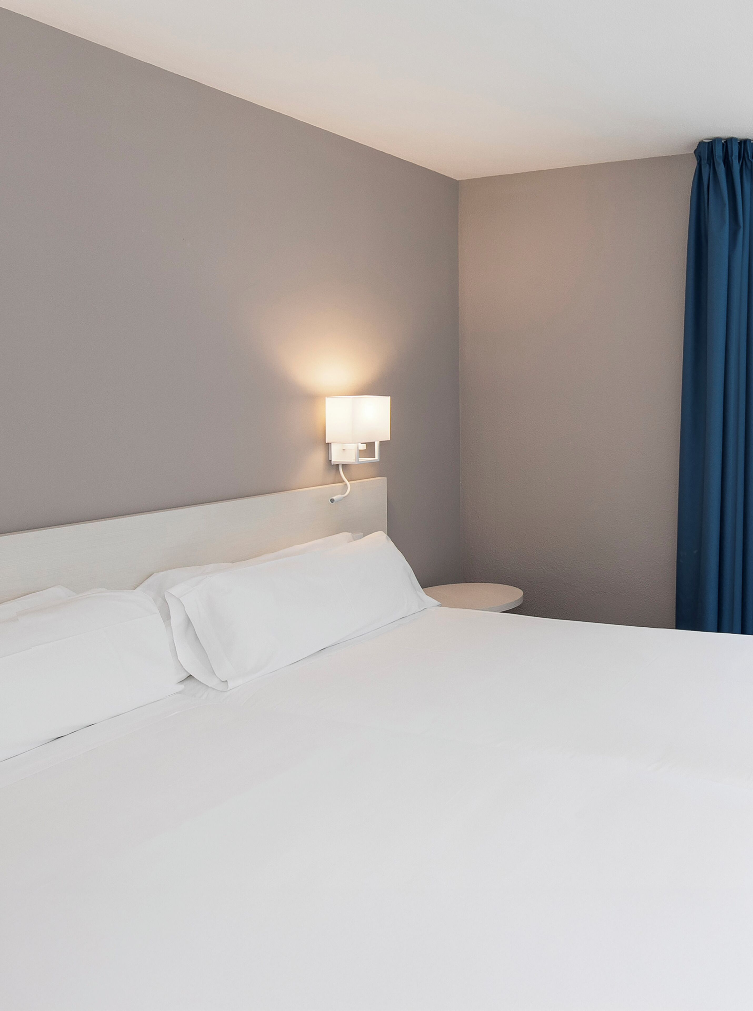 Une chambre d’hôtel avec un lit, une table de chevet, une lampe fixée au mur et une fenêtre avec un rideau.