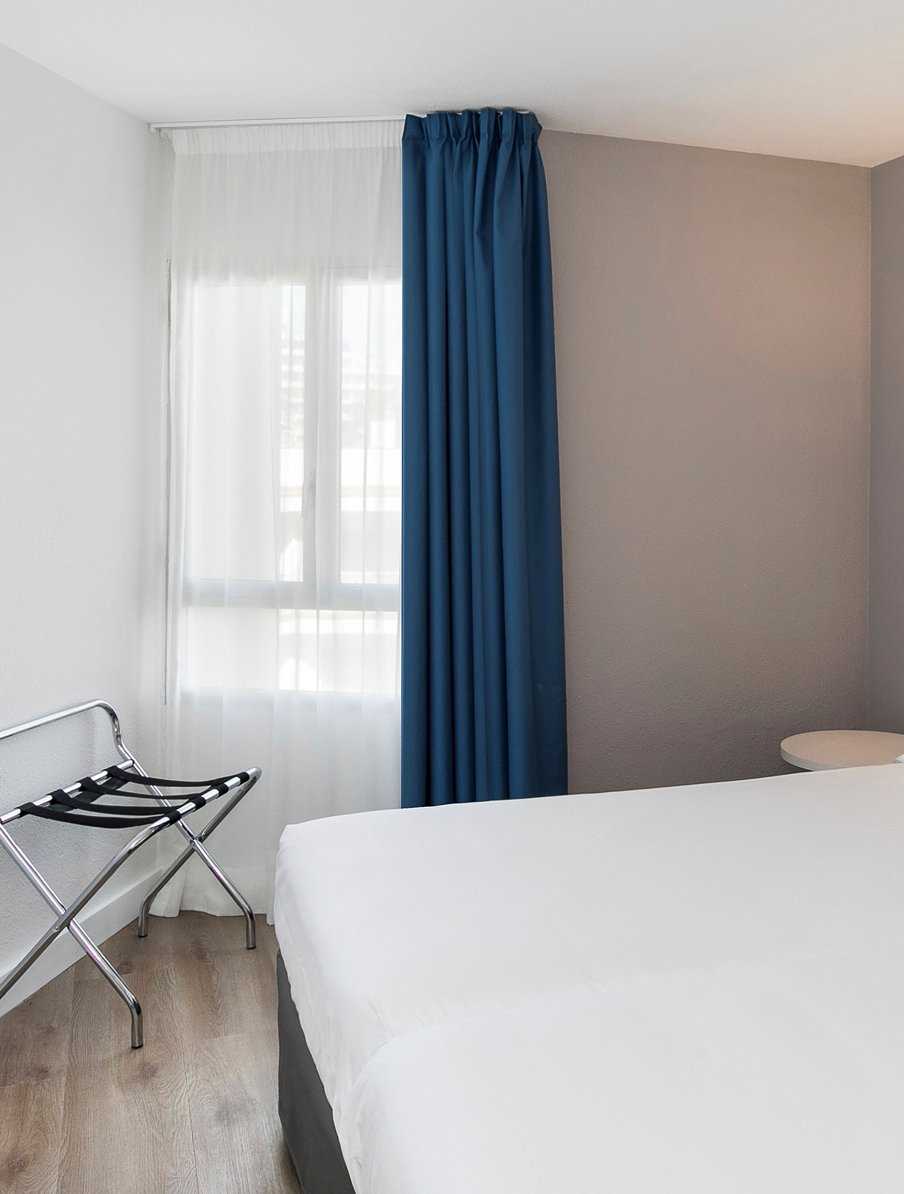 Une chambre d’hôtel avec un lit, un étendoir métallique, une fenêtre avec des rideaux légers et un rideau bleu uni.