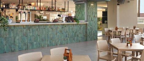 A bar area with a teal-colored counter, a laptop, and a coffee machine. There are several tables and chairs in the seating area.
