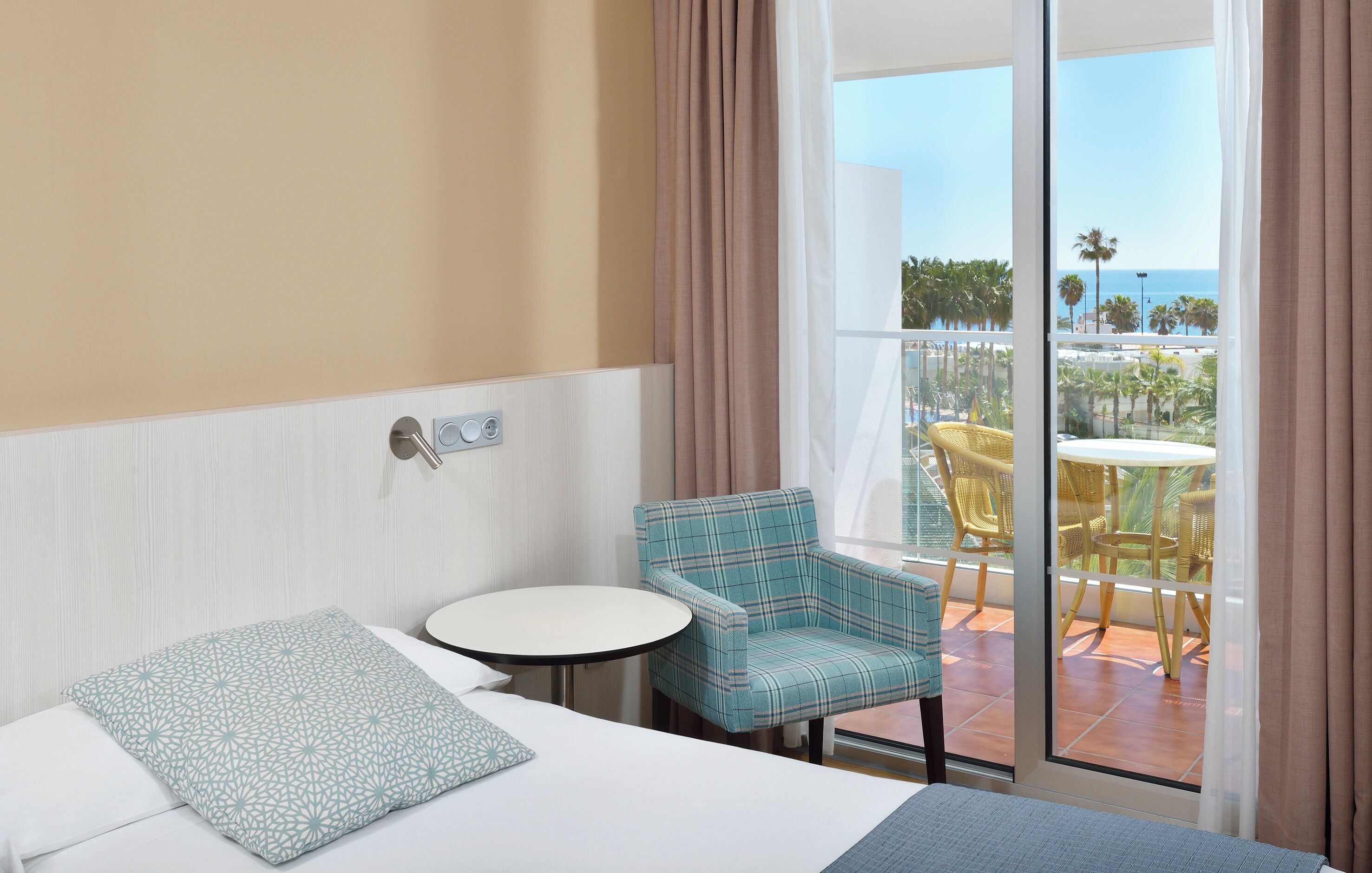 Een hotelkamer met een bed, een klein rond tafeltje, een geruit fauteuil en uitzicht op een balkon met buitenmeubilair.