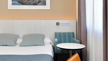 A hotel room with a bed, a seating area with a table, a chair, and a wall-mounted power outlet.