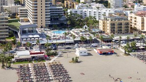 Luftaufnahme einer Küstenstadt mit Strand, Hotels und einem Bergpanorama im Hintergrund.
