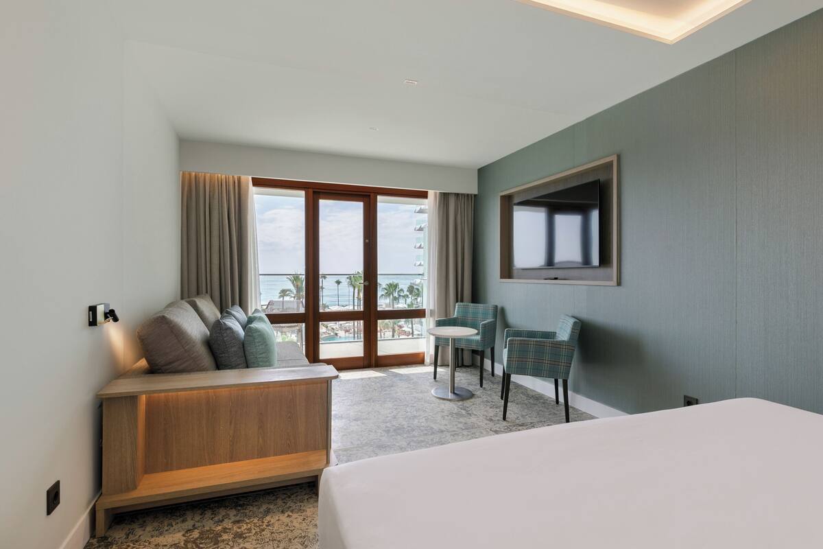 Een hotelkamer met een bed, een zithoek met een bank en stoelen, een aan de muur bevestigde televisie en een groot raam met uitzicht op de oceaan.