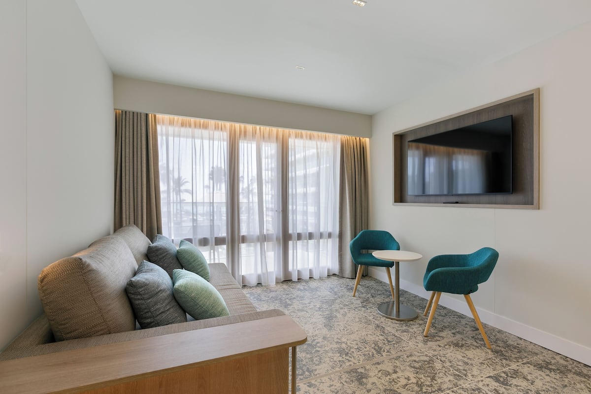 Een moderne hotelkamer met een sofa, twee stoelen, een klein tafeltje, een flatscreen televisie en grote ramen met dunne gordijnen.