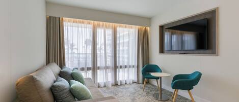 A modern hotel room with a sofa, two chairs, a small table, a flat-screen TV, and large windows with sheer curtains.