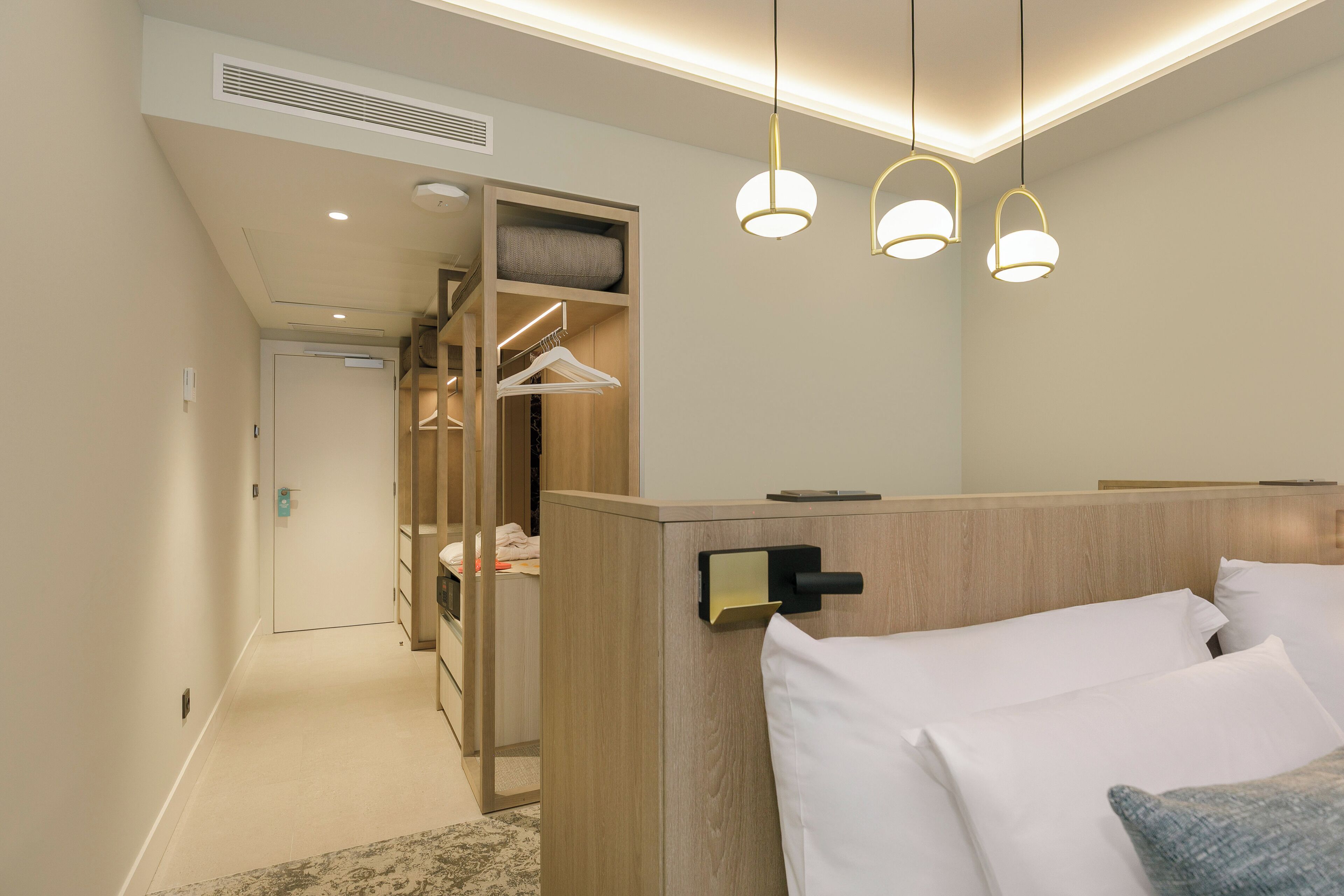 Une chambre d’hôtel avec une tête de lit en bois, un lit avec du linge de maison blanc, un dressing avec des vêtements suspendus et des lampes suspendues.