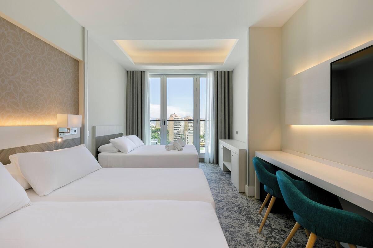 Een hotelkamer met twee bedden, een bureau, een stoel, een televisie en een groot raam met uitzicht op gebouwen.