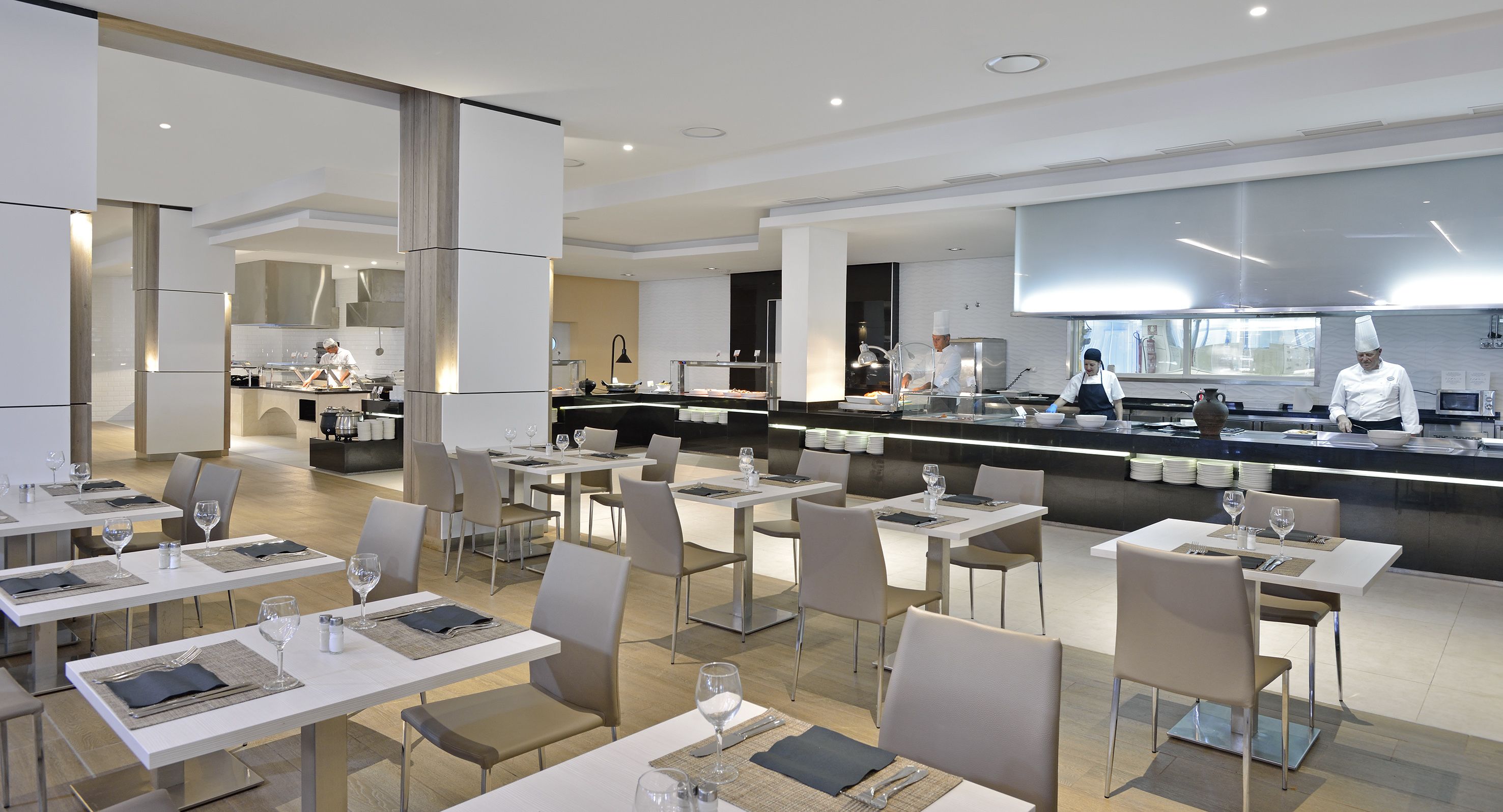 A modern restaurant with white tables and chairs, a kitchen area, and a bar.