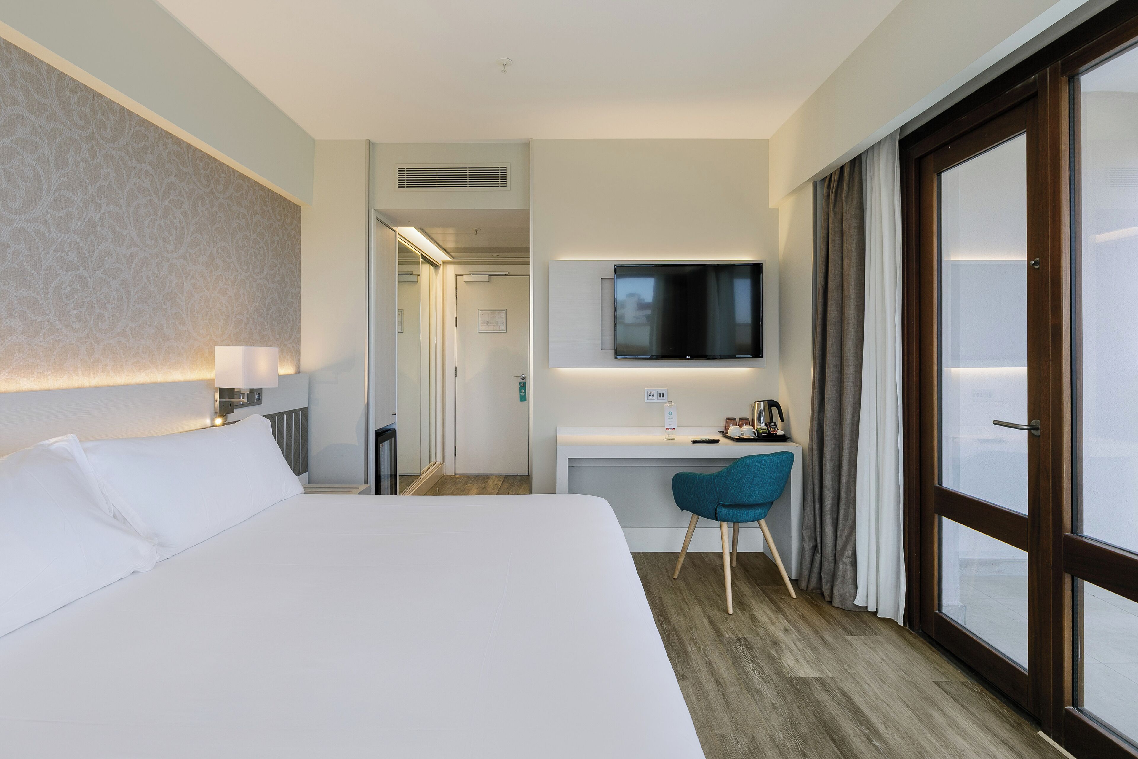 Una habitación de hotel con una cama grande, un escritorio, un televisor de pantalla plana, una silla y un amplio ventanal con cortinas.