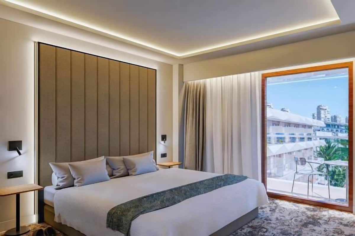 A hotel room with a large bed, a view of the city, and a balcony with a chair.