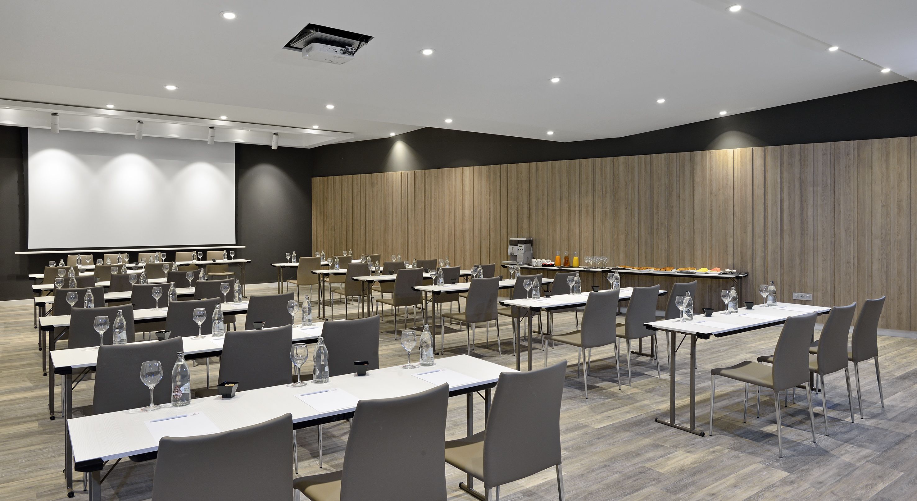 Uma sala de conferências com várias mesas, cadeiras, tela de projeção e um espaço para café.