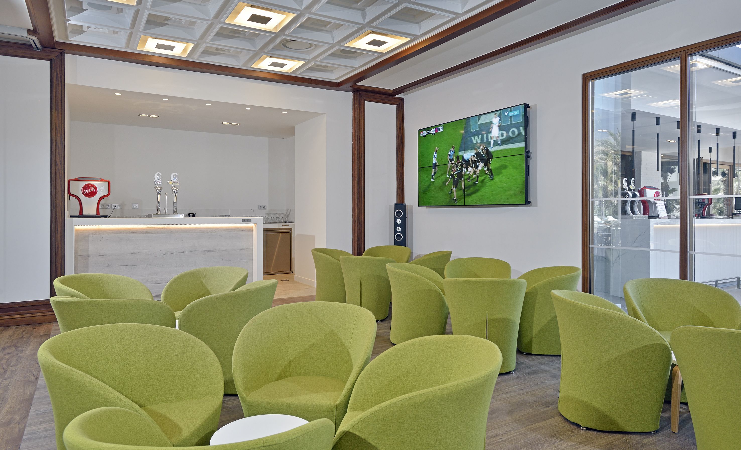 A modern hotel lobby with green chairs arranged in rows, a reception desk with a Coca-Cola machine, and a television screen displaying a rugby match.