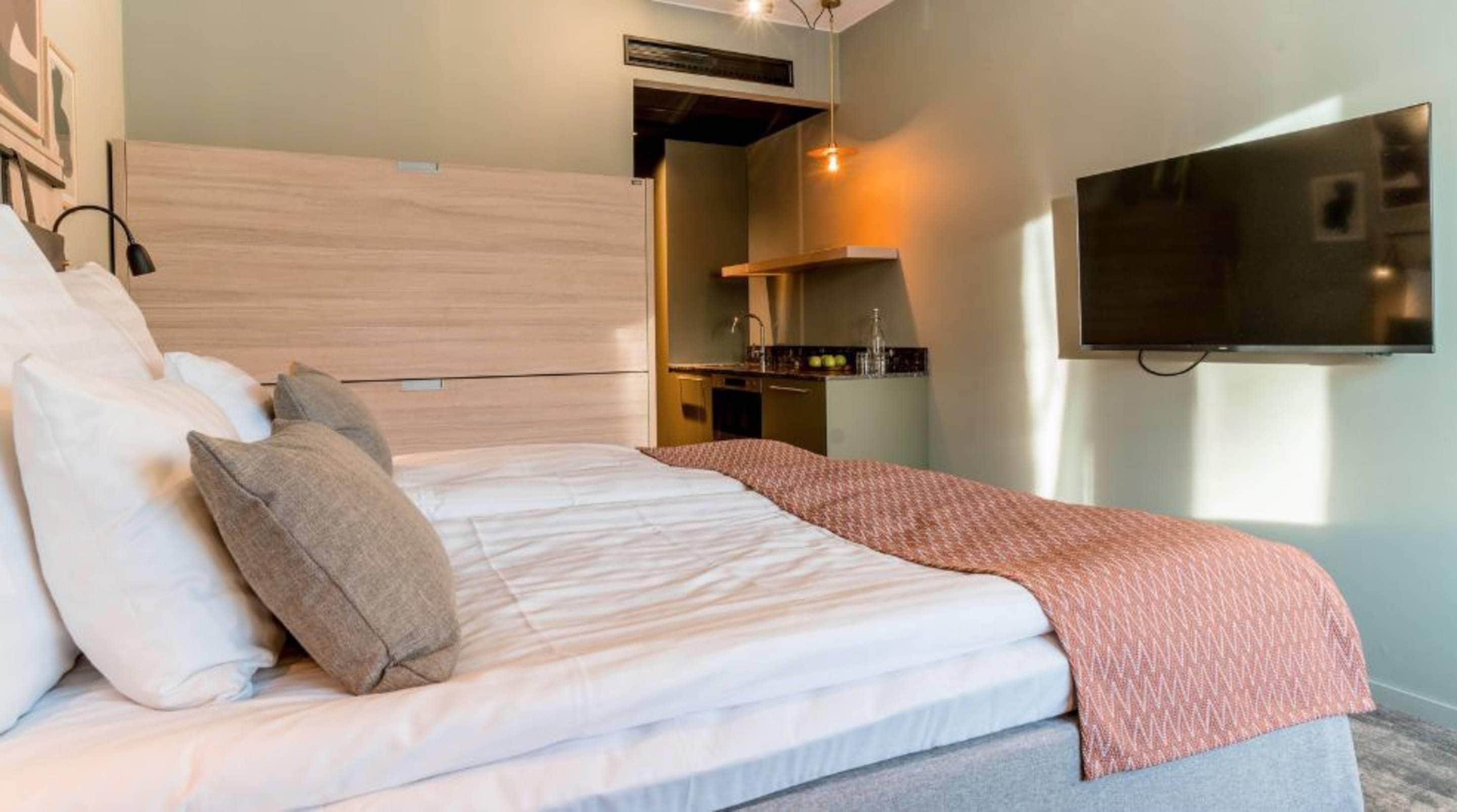 A hotel room with a bed, a TV mounted on the wall, and a kitchenette area.
