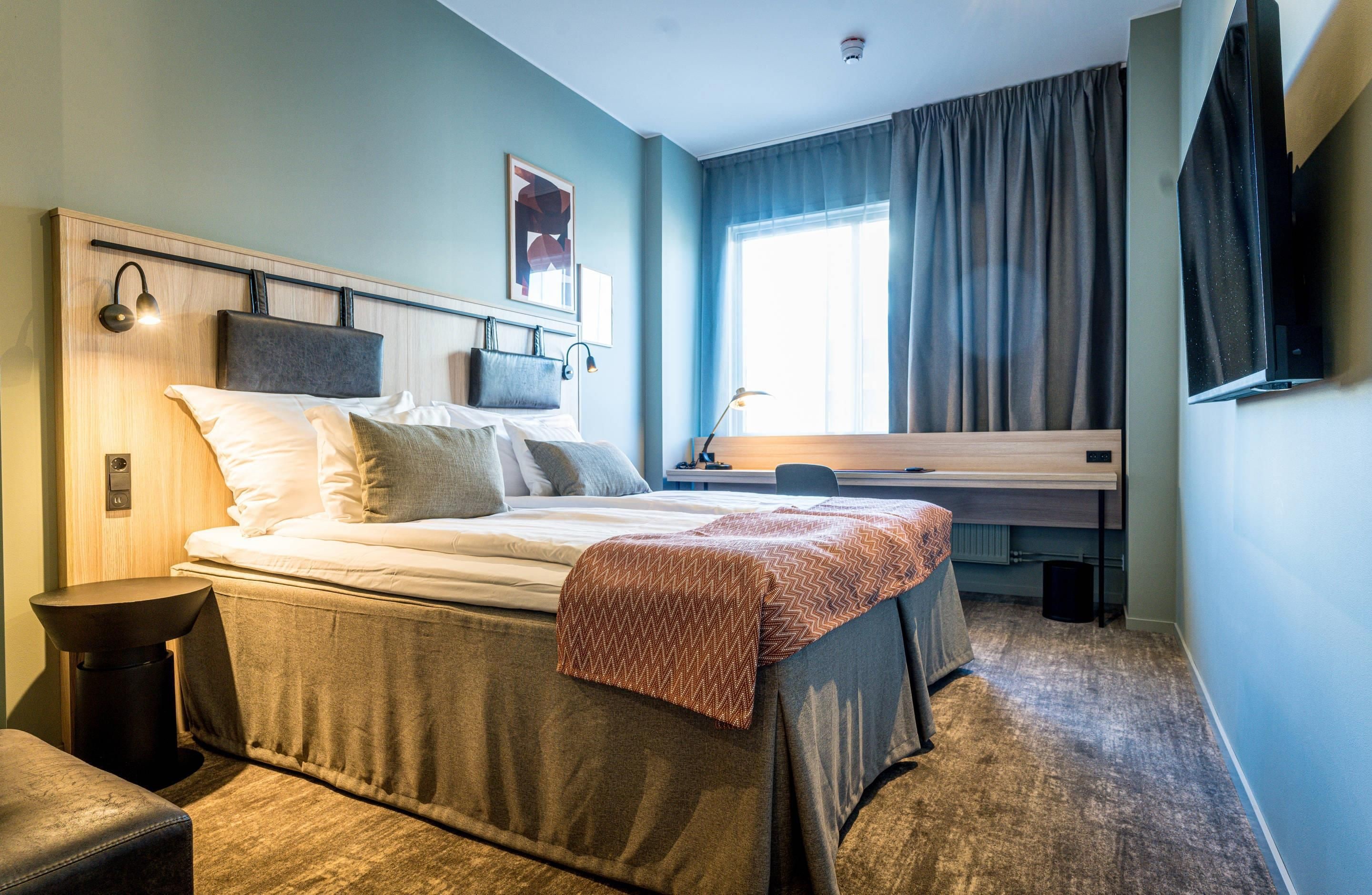 A hotel room with a bed, bedside table, lamp, and a desk with a chair.