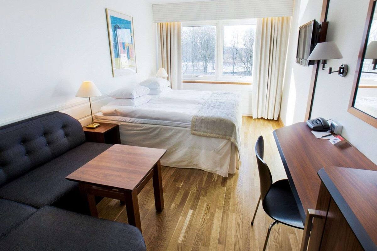 A hotel room with a bed, a sofa, a desk, a chair, a lamp, a phone, and a painting on the wall.