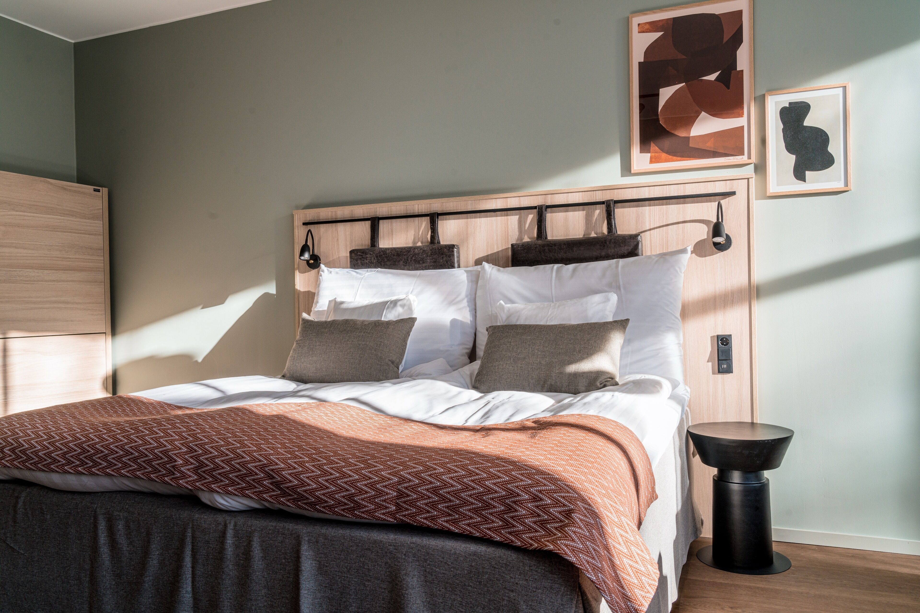A neatly made bed with a patterned blanket, a bedside table, and two abstract artworks on the wall.