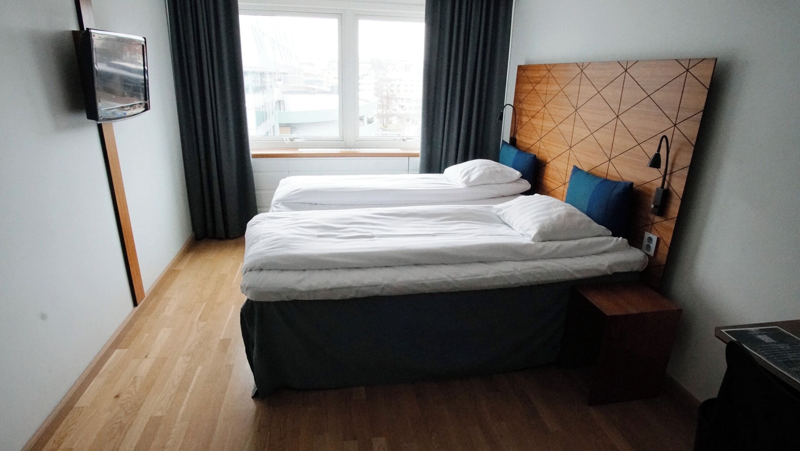 A double bed with white linens, a wooden headboard with a geometric pattern, a bedside table, and a window with curtains.