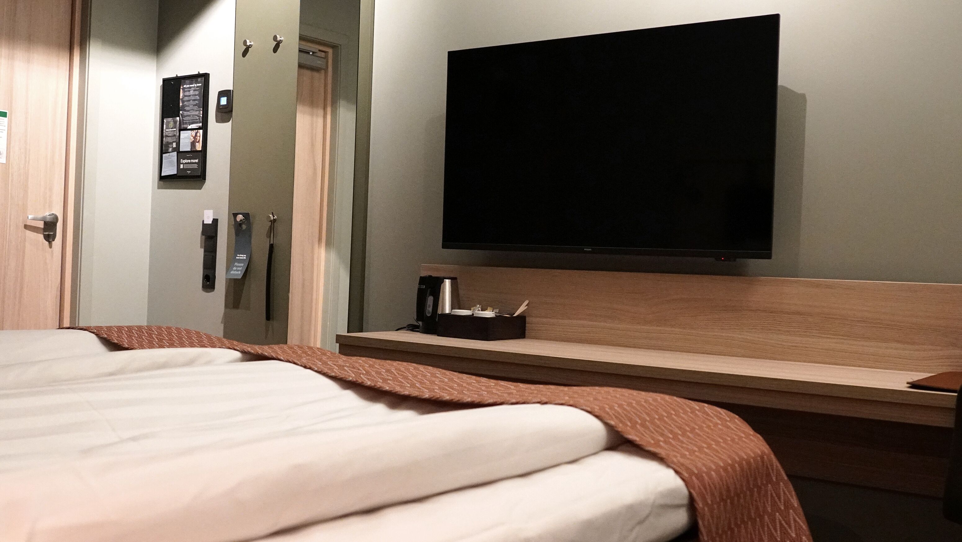 Habitación de hotel con cama, televisor de pantalla plana montado en la pared, mesita de noche con taza y control remoto, y puerta con cerradura y lector de tarjetas.