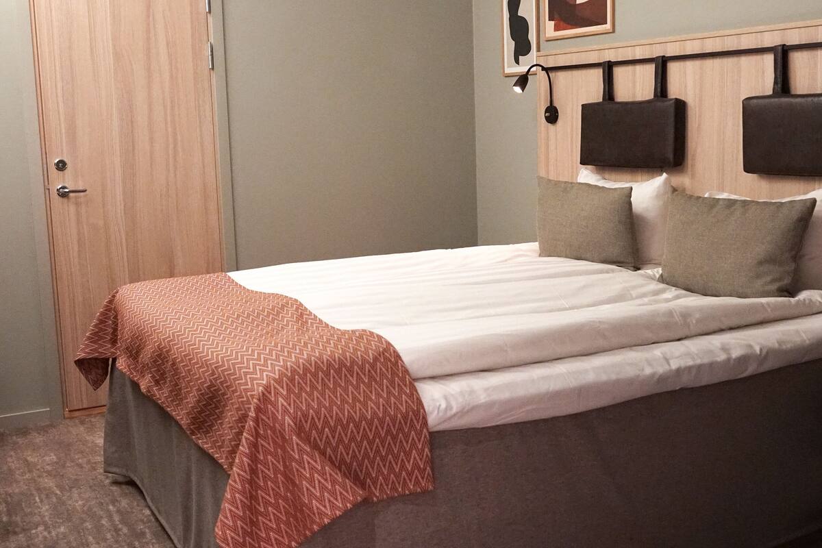A neatly made bed with a patterned throw, a headboard with two wall-mounted lamps, and a wooden door.
