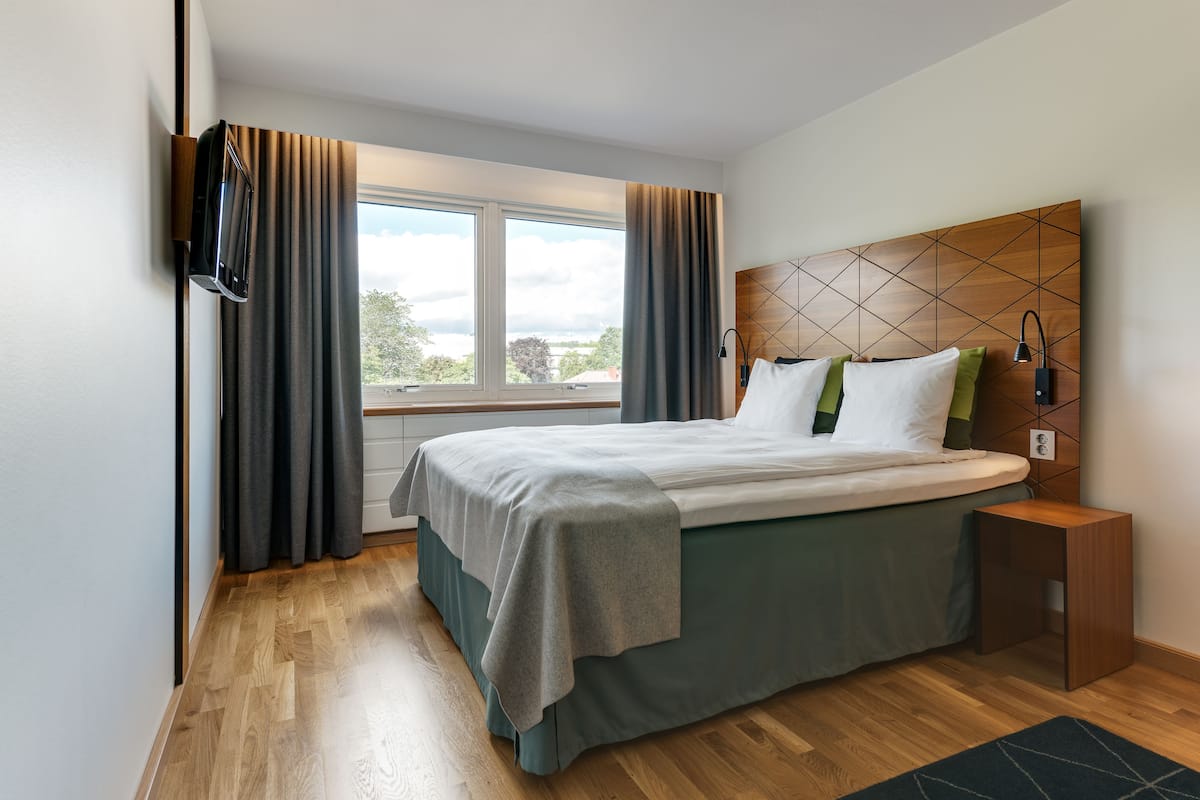 A modern hotel room with a large bed, a wooden headboard, a nightstand, and a view of the outdoors.