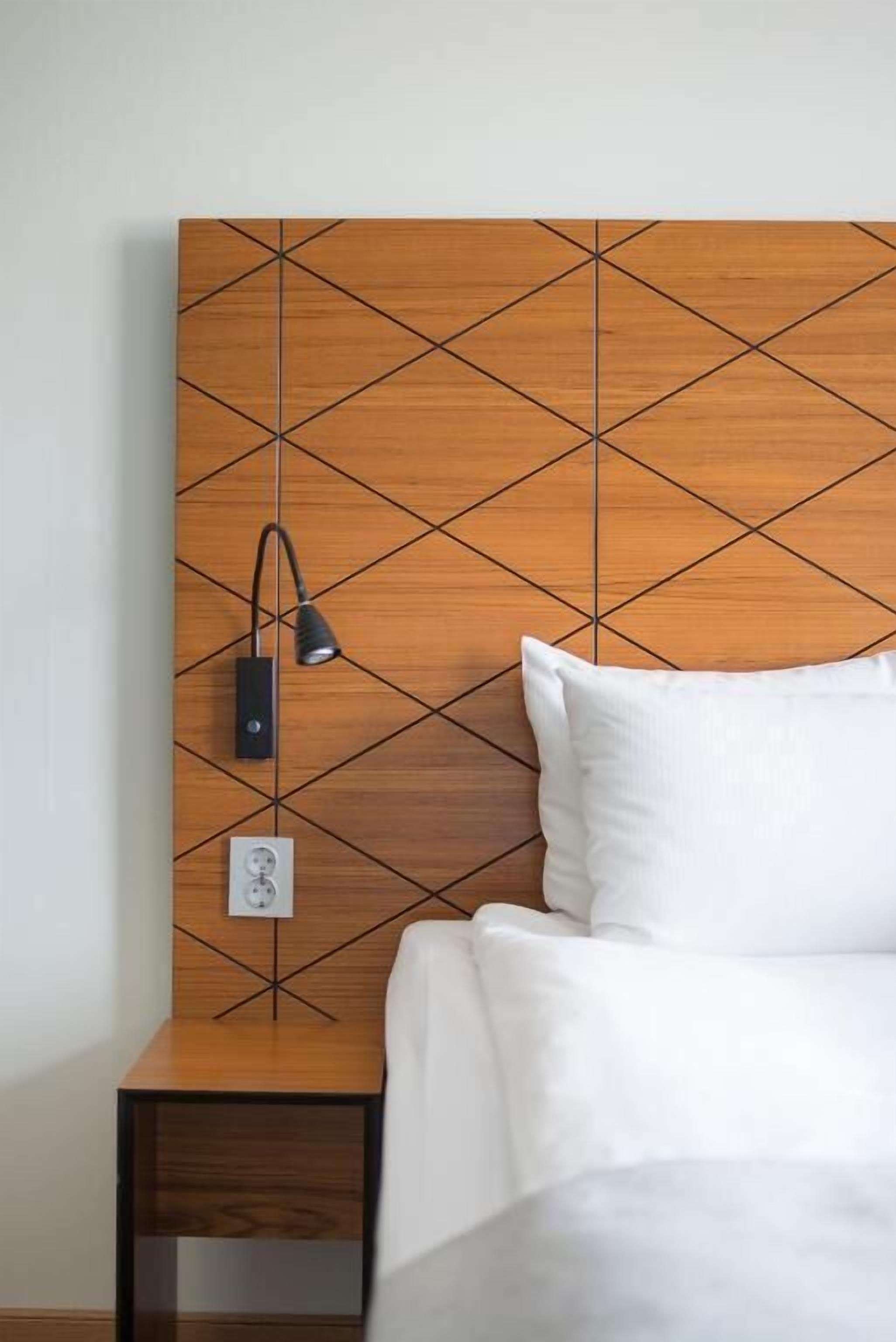 Un lit bien fait, avec du linge de lit blanc, une tête de lit en bois ornée d’un motif géométrique, une lampe de chevet et une prise électrique sur le mur.
