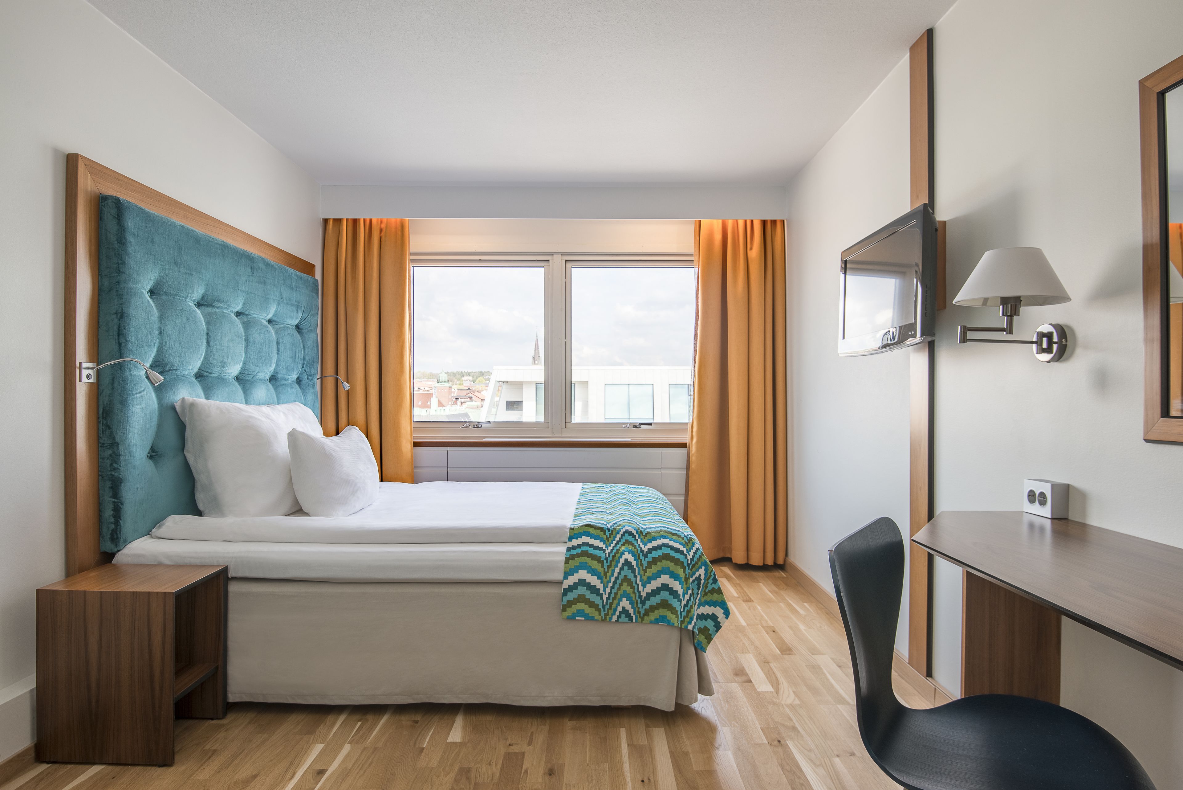 A modern hotel room with a bed, a desk, a chair, a TV, and a window with curtains.
