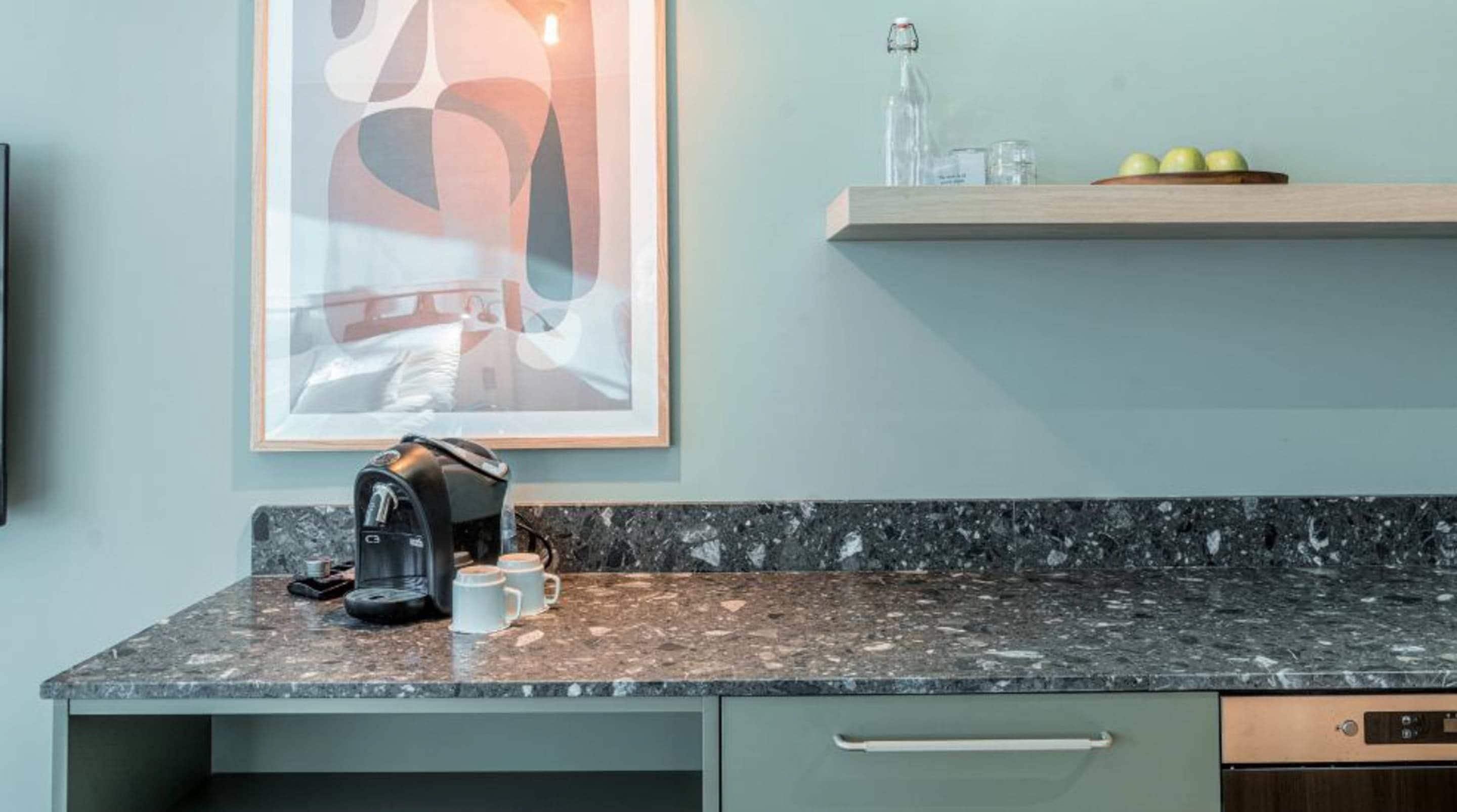 A kitchen with a coffee maker, a framed abstract artwork, and a fruit bowl on a shelf.