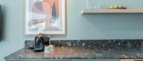 A kitchen with a coffee maker, a framed abstract artwork, and a fruit bowl on a shelf.