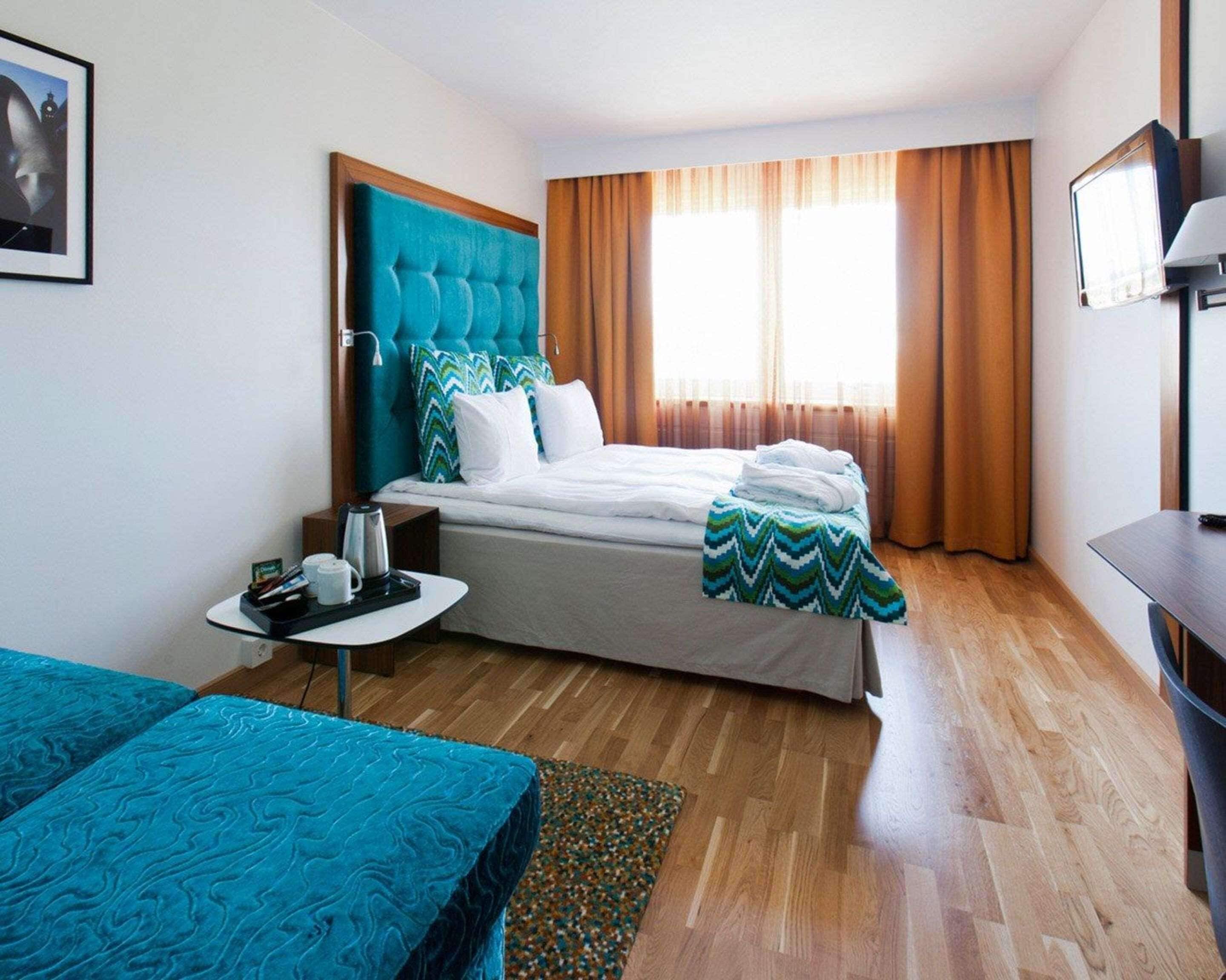 A hotel room with a bed, a small table with a kettle, a TV, and a window with curtains.