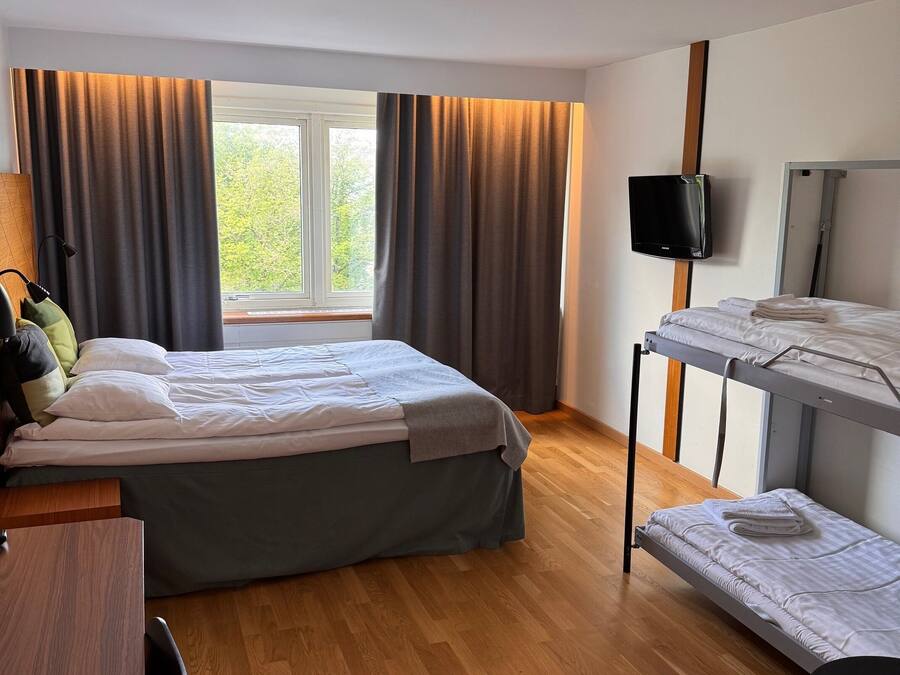 A hotel room with a bed, a desk, a bunk bed, a TV, and a window with curtains.