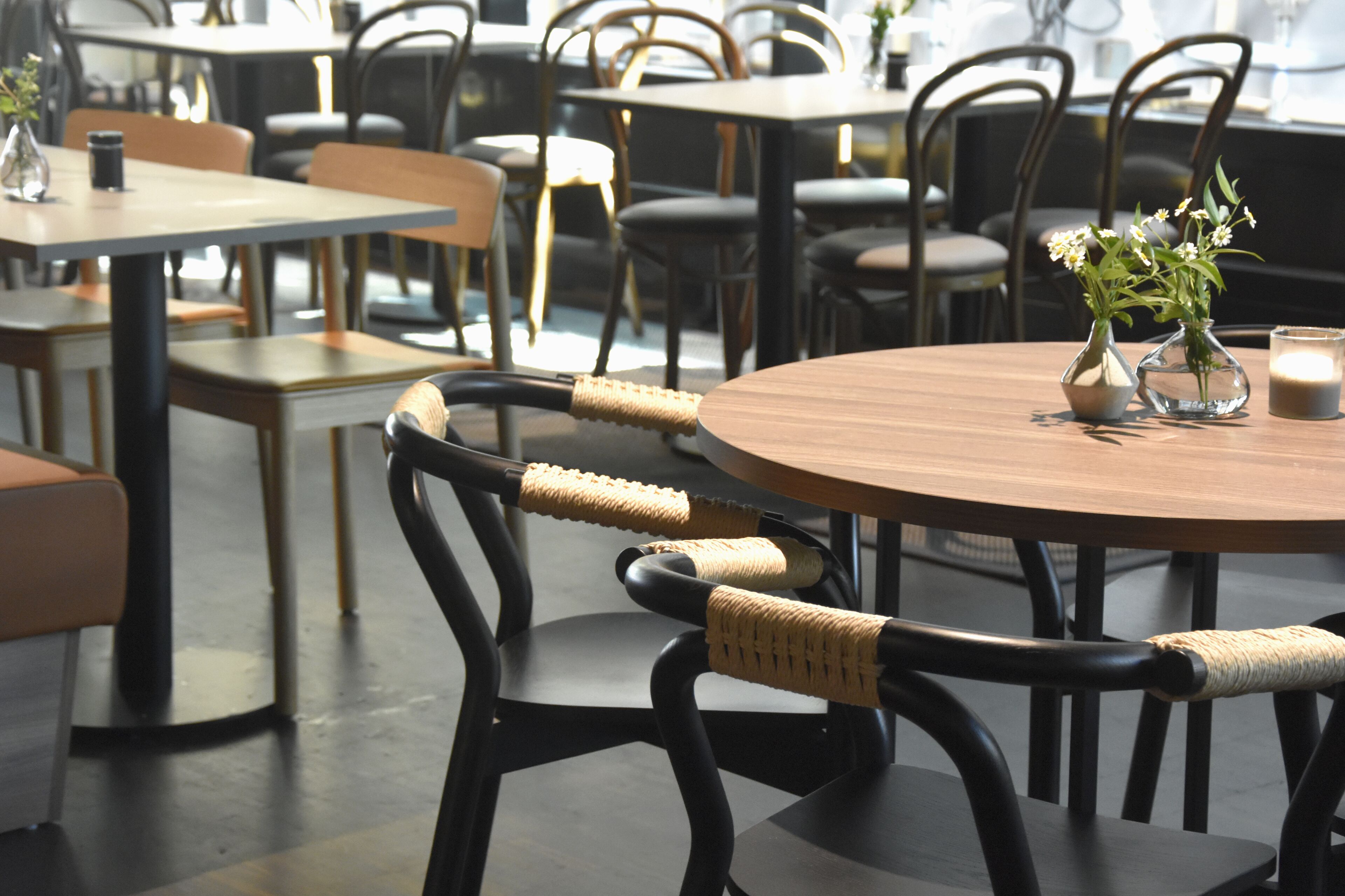 Interior de restaurante con mesas redondas, sillas negras con reposabrazos trenzados y pequeños jarrones con flores.