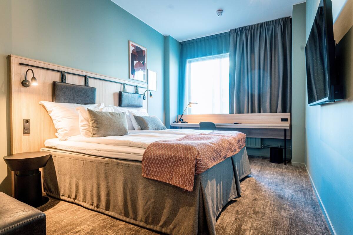 A hotel room with a bed, bedside table, lamp, and a TV mounted on the wall.