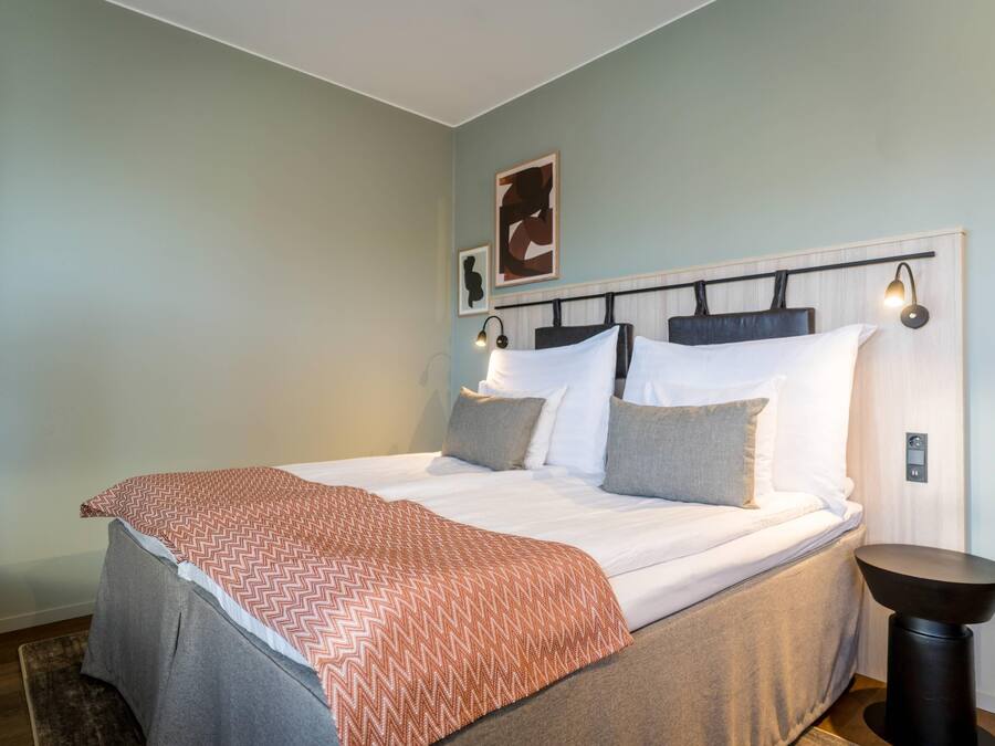 Un dormitorio con cama, almohadas, una mesita de noche y una lámpara de pared.