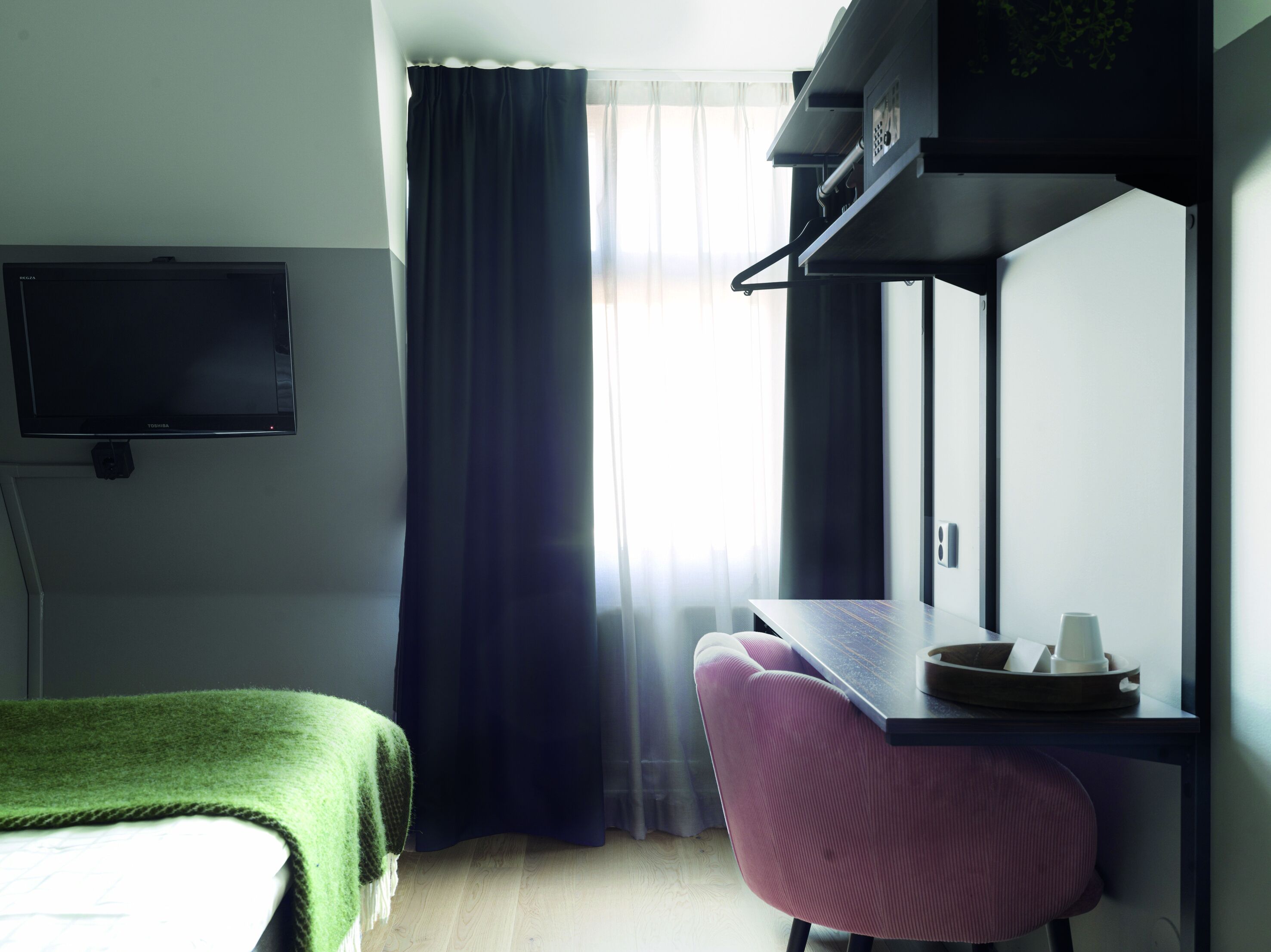 Ein Zimmer mit einem Bett, einem Schreibtisch mit Stuhl, einem an der Wand montierten Fernseher und einem Fenster mit Vorhängen.