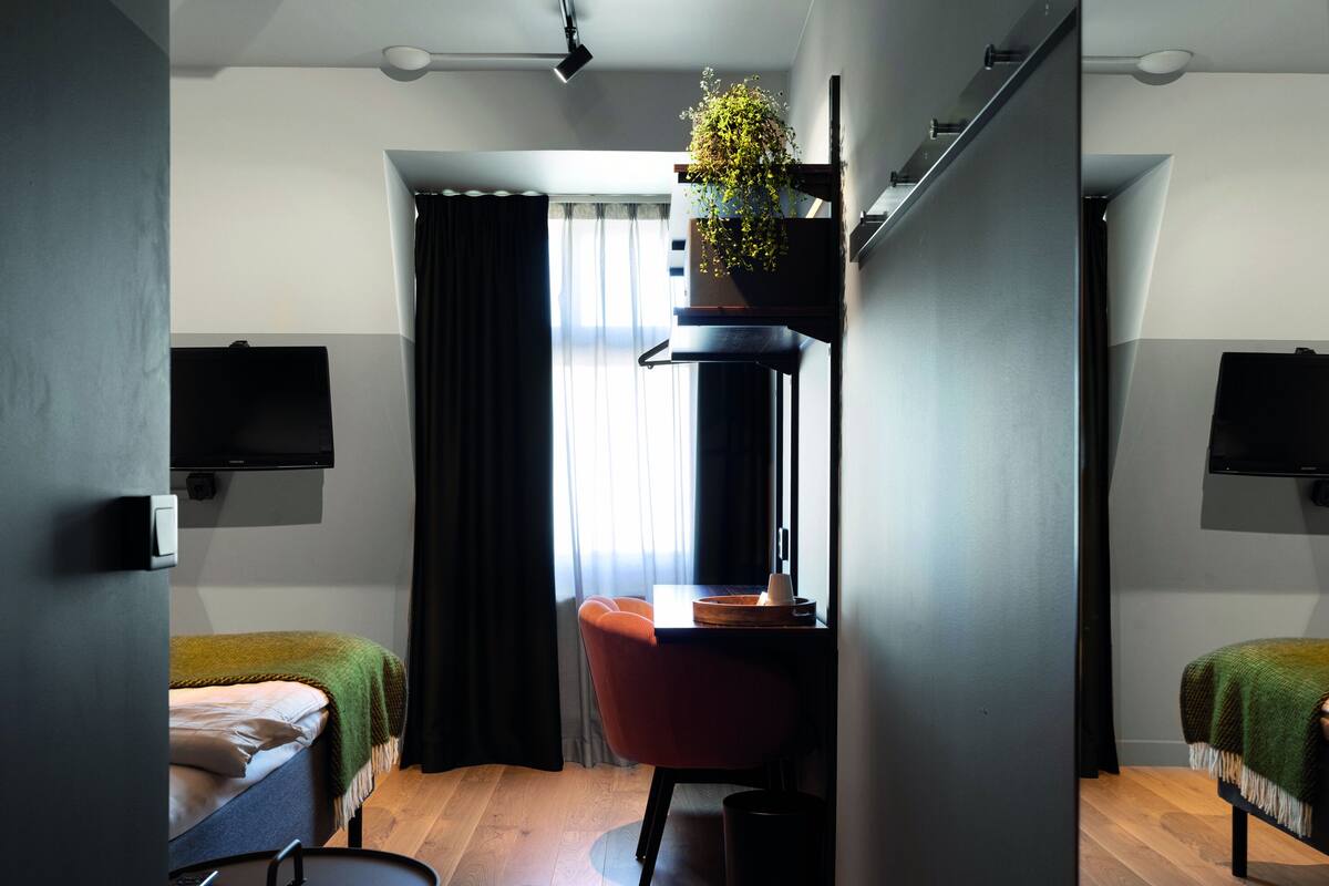 Ein kompaktes Hotelzimmer mit Bett, Schreibtisch mit Stuhl, einem kleinen Tisch, einer Topfpflanze und einem an der Wand befestigten Fernseher.