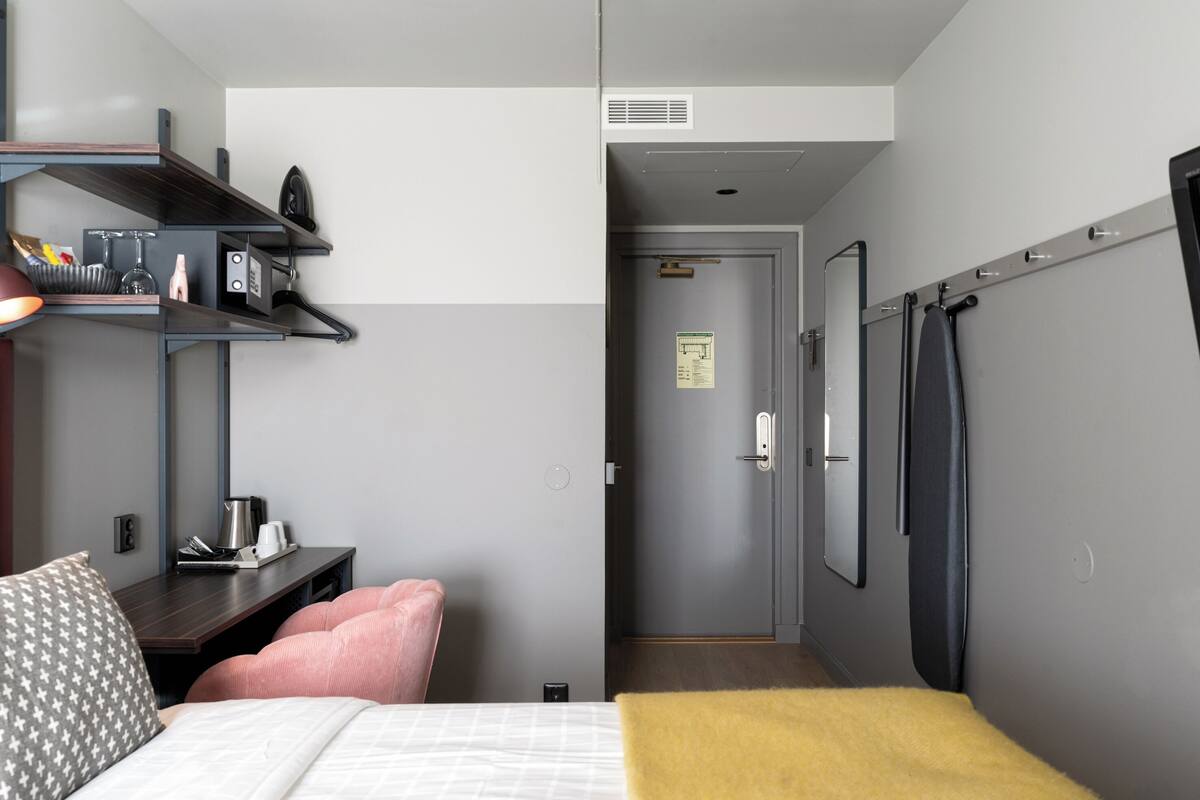 Ein Hotelzimmer mit einem Bett, einem Schreibtisch mit Stuhl, einem Regal mit diversen Gegenständen und einer Tür zu einem anderen Raum.