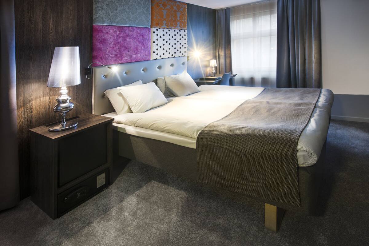 Ein Hotelzimmer mit einem Bett, zwei Nachttischen mit Lampen, einem Beistelltisch, einer Wand mit gemustertem Tapeten und einem Fenster mit Vorhängen.