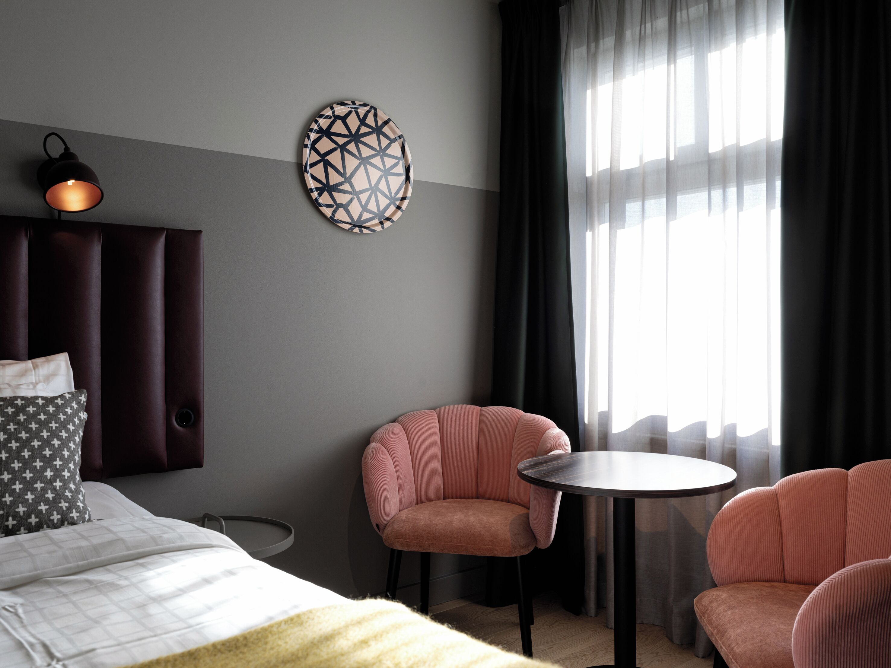 A modern hotel room with a bed, two pink upholstered chairs, a small round table, a wall-mounted lamp, and a geometric wall art piece.